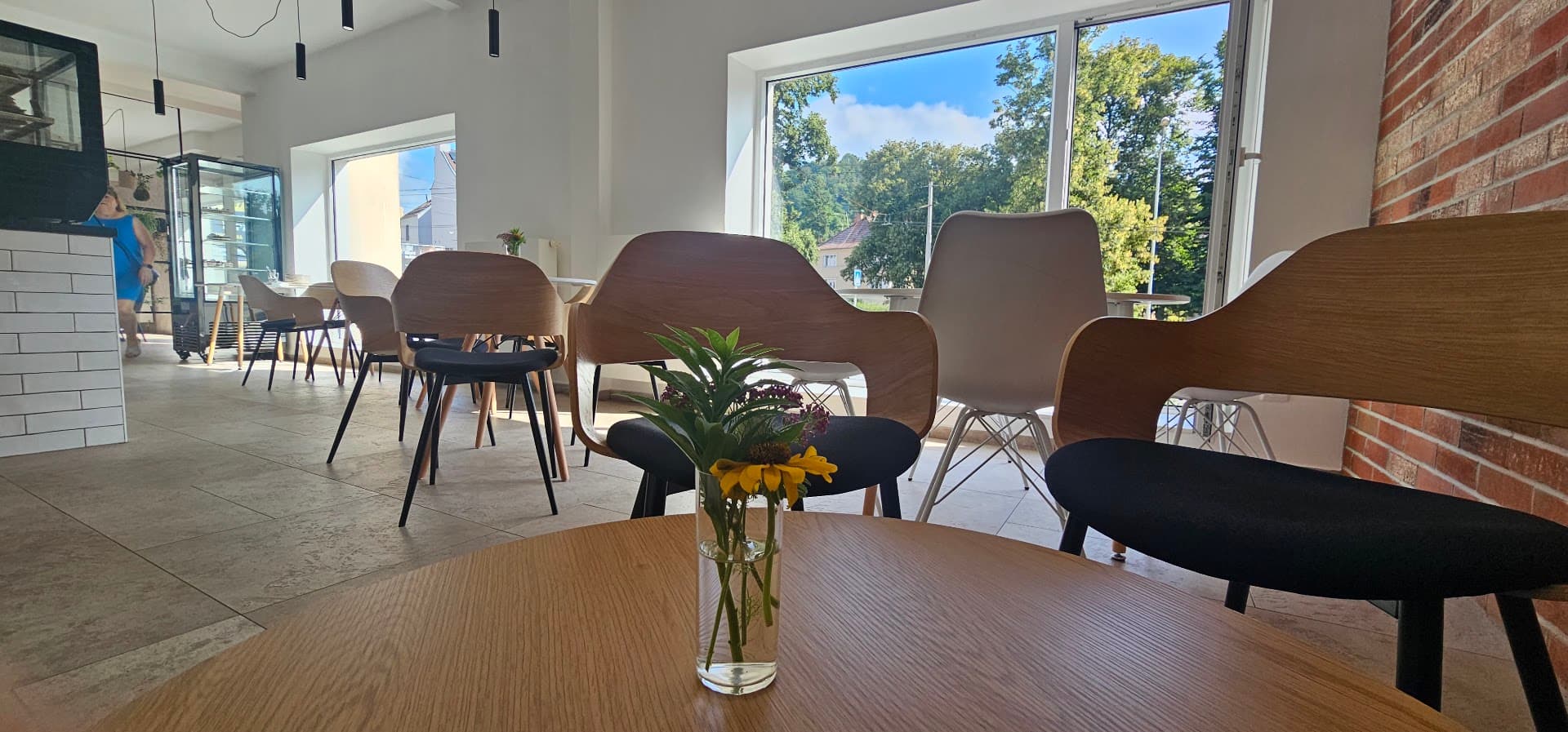 Prosluněný interiér Nina Kafé — dřevěné židle, cihlová stěna a čerstvé květiny na stolech